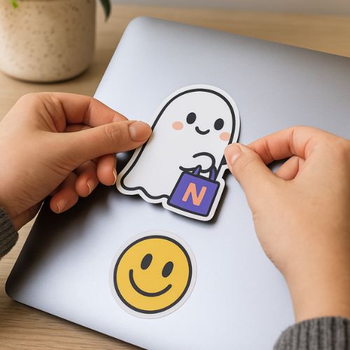 Two hands are placing a cute ghost sticker holding a shopping bag with the letter N onto a laptop, which already has a yellow smiley face sticker on it. A potted plant is visible in the background.
